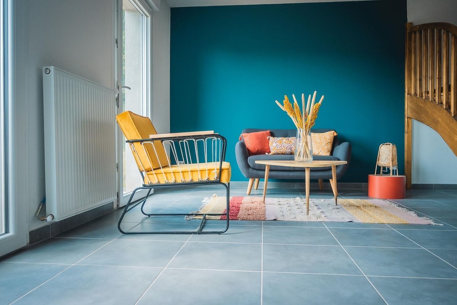 Cozy living room with teal wall, yellow chair, sofa, coffee table, vase, and rug on blue tile floor