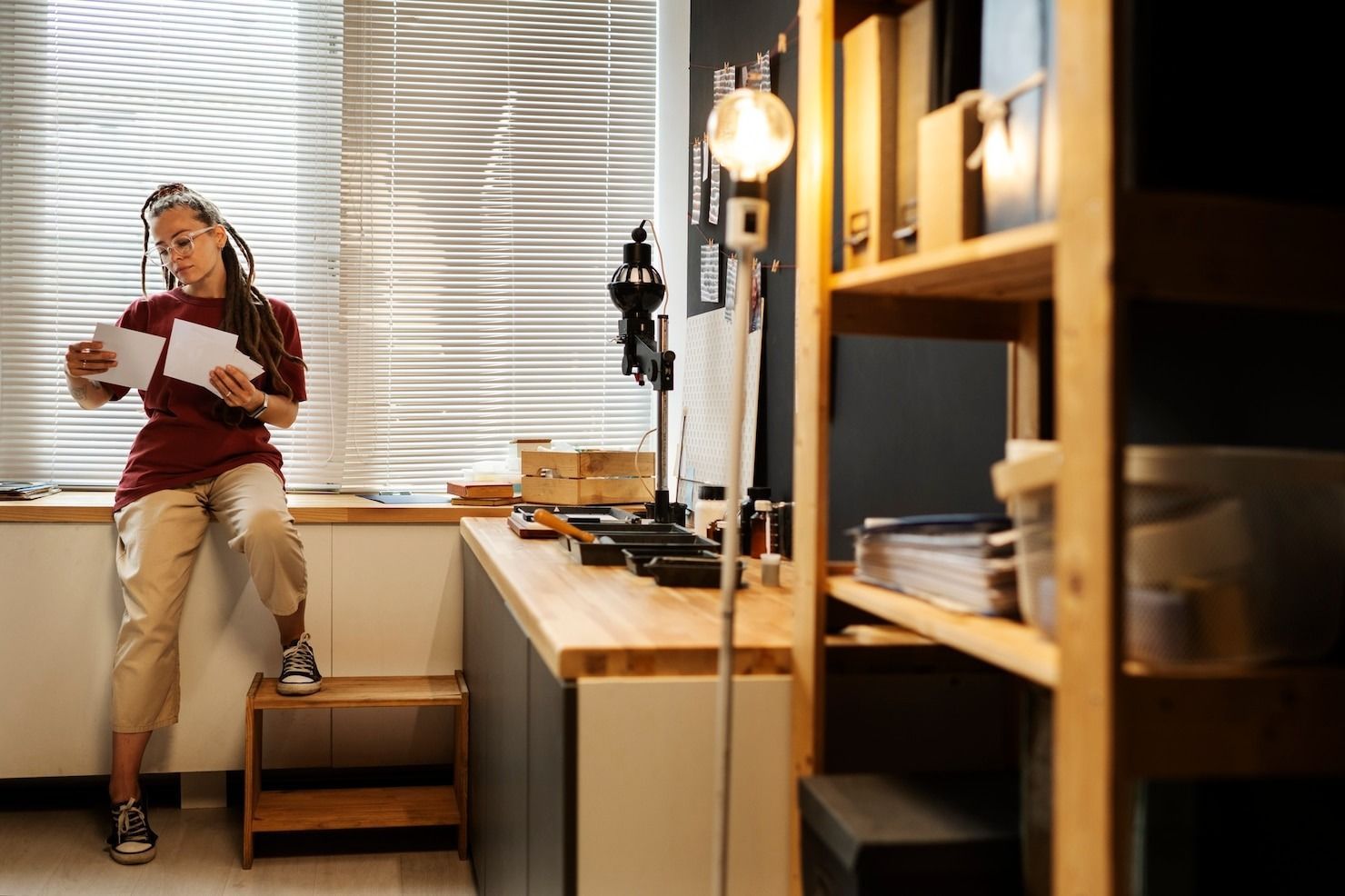 Person reading a book in a cozy room with shelves, a lamp, and a desk by the window.