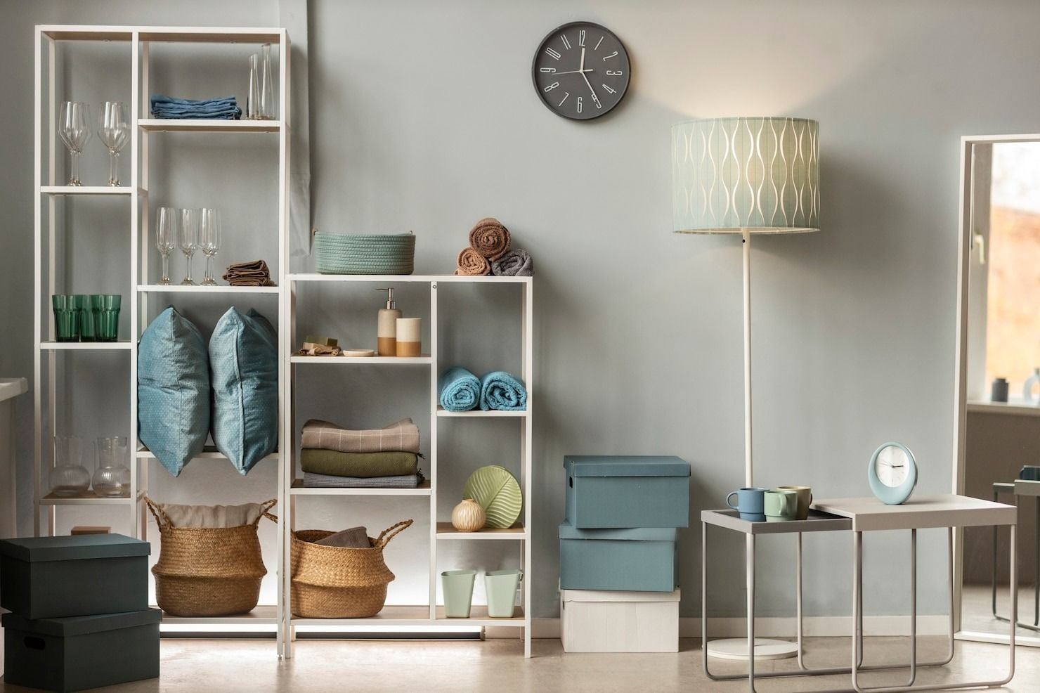 Minimalist living room with white shelving, blue storage bins, baskets, lamp, wall clock, and small table