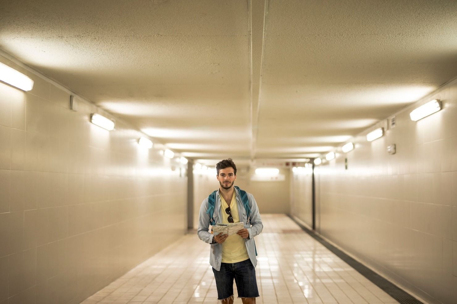 Man standing in a tiled pedestrian tunnel under bright lights, holding a bottle and looking at the camera.