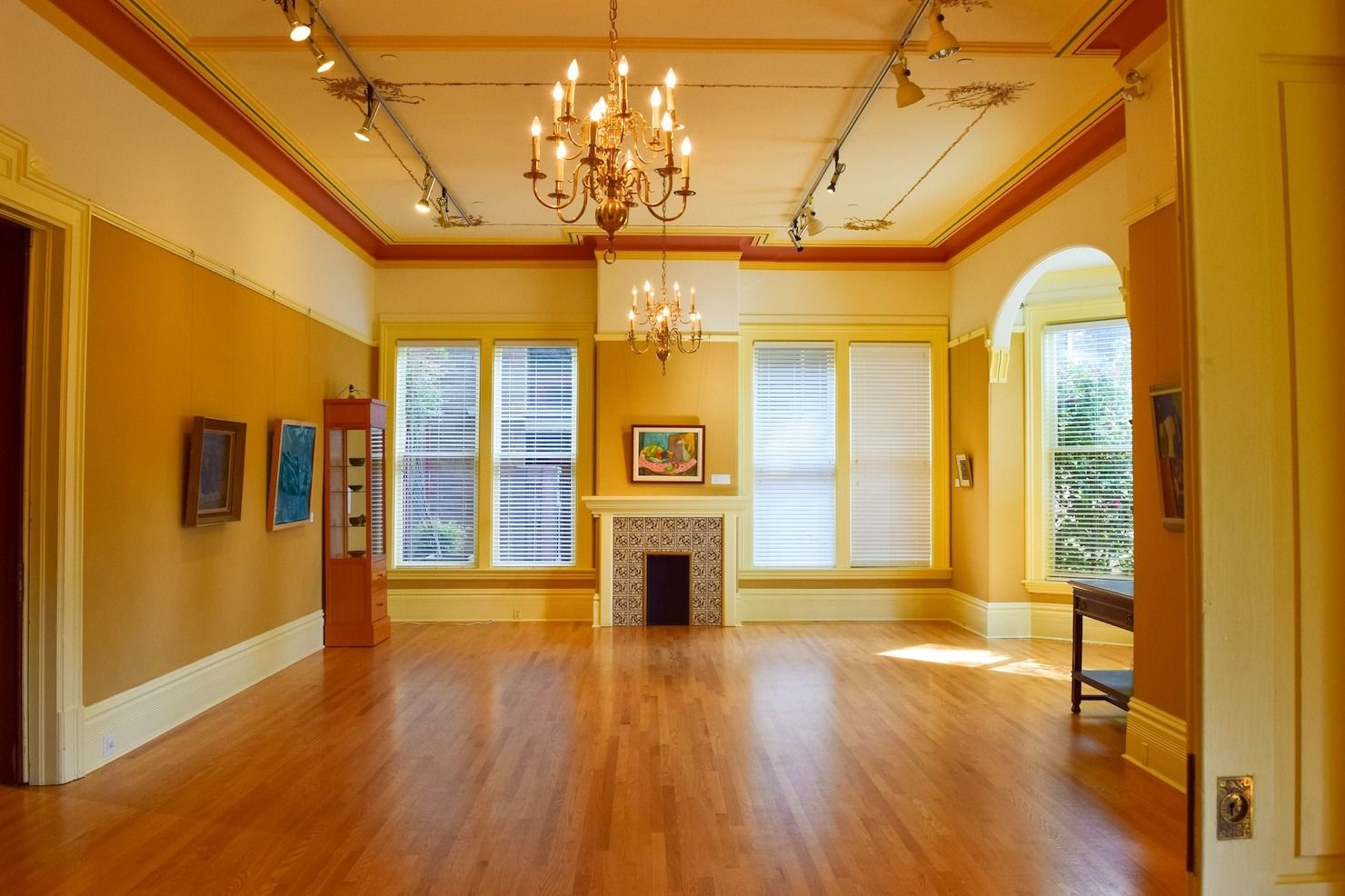 Empty yellow room with hardwood floors, chandelier, fireplace, and tall windows overlooking greenery