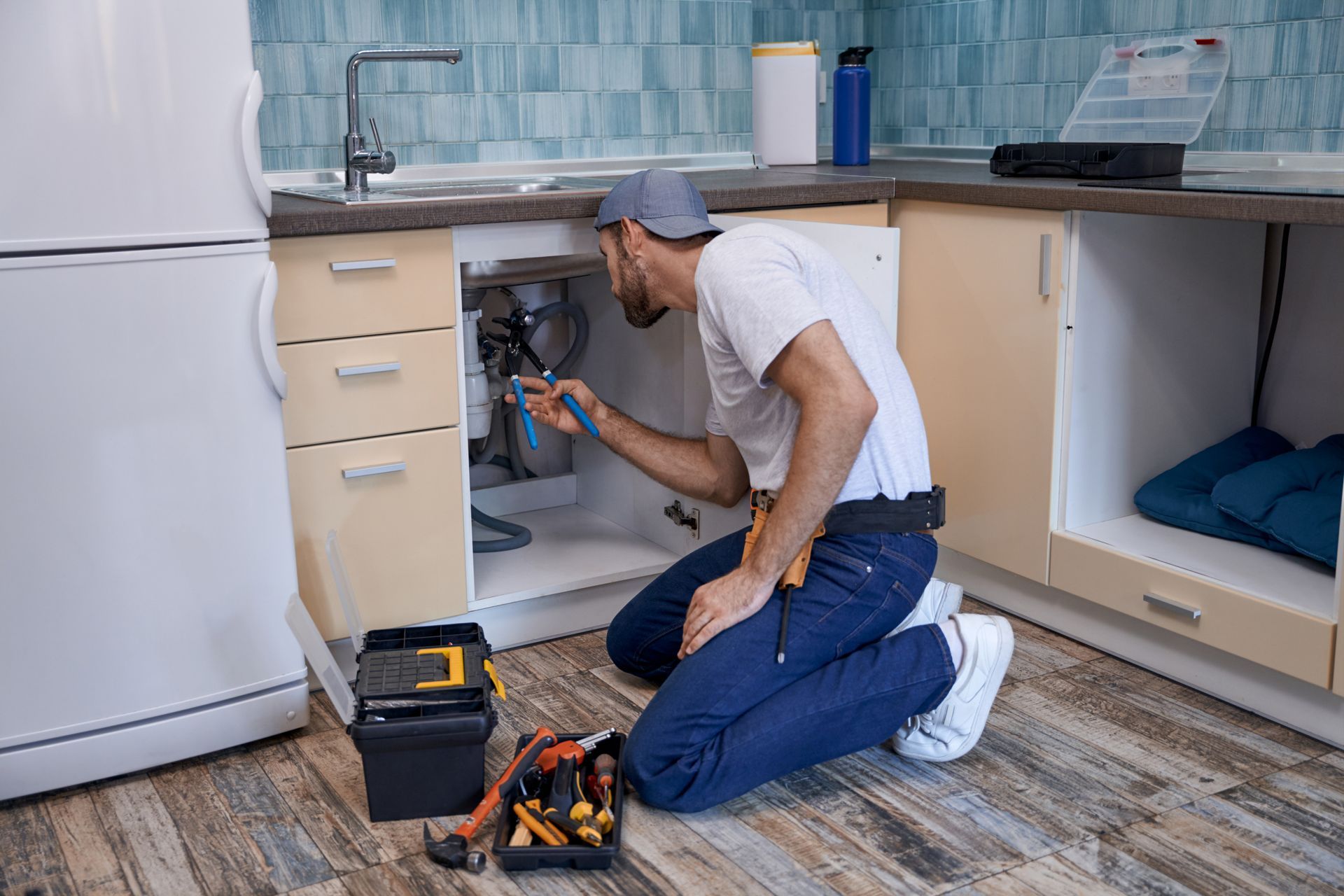 Plumber kneeling under kitchen sink, repairing pipes with tools on the floor.