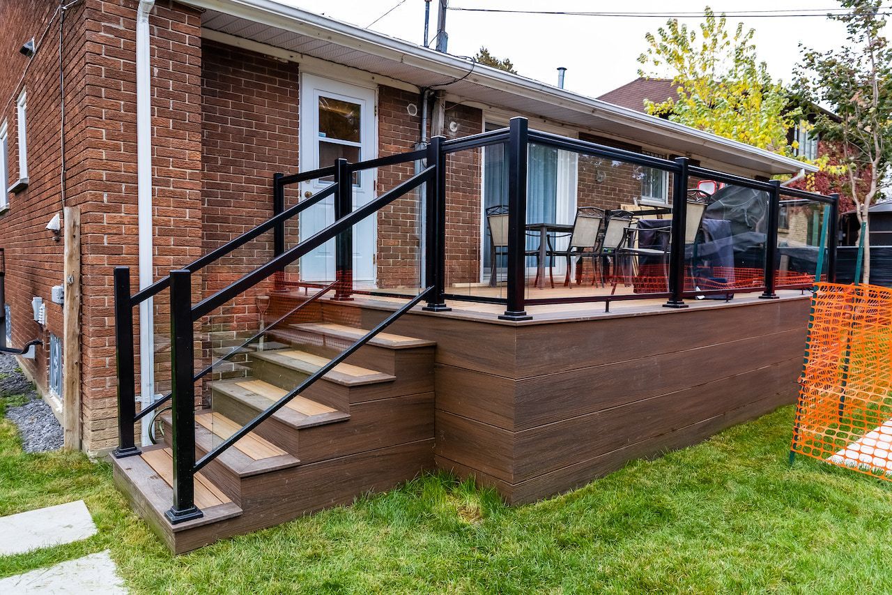 Maison en briques avec terrasse et balustrade en verre, escalier en bois et pelouse verdoyante.