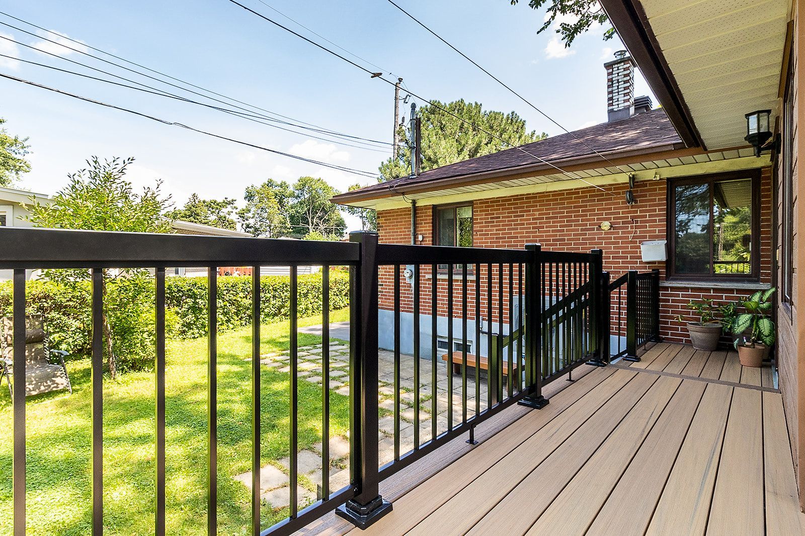 Terrasse en bois avec rambarde noire, donnant sur une cour gazonnée avec une maison en briques en arrière-plan.