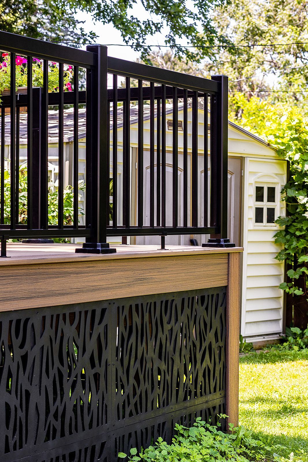 Rambarde noire avec panneau inférieur décoratif sur une terrasse en bois devant un abri de jardin blanc.
