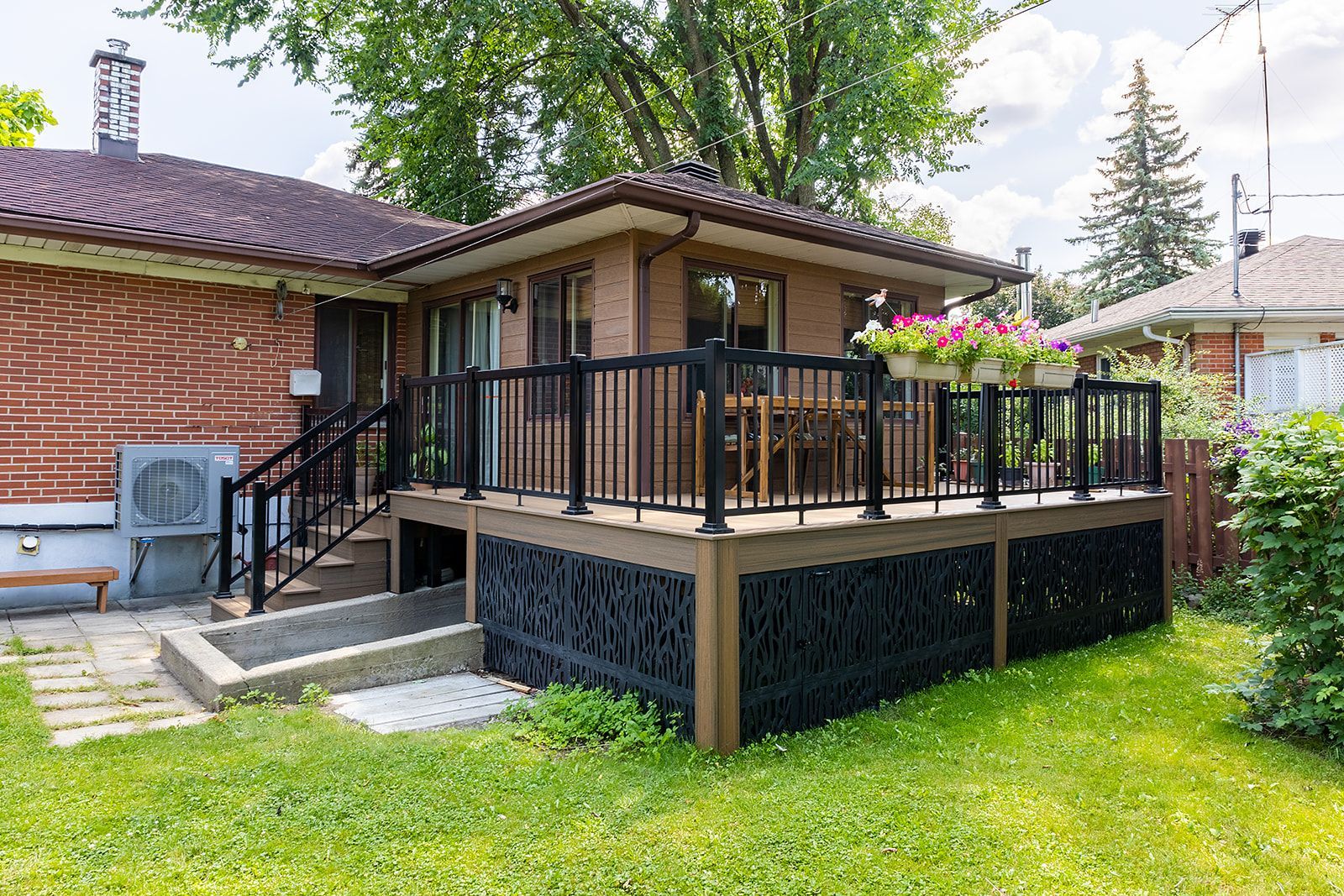 Terrasse arrière avec balustrade noire et lambris décoratifs. Maison en briques rouges, pelouse verte et journée ensoleillée.