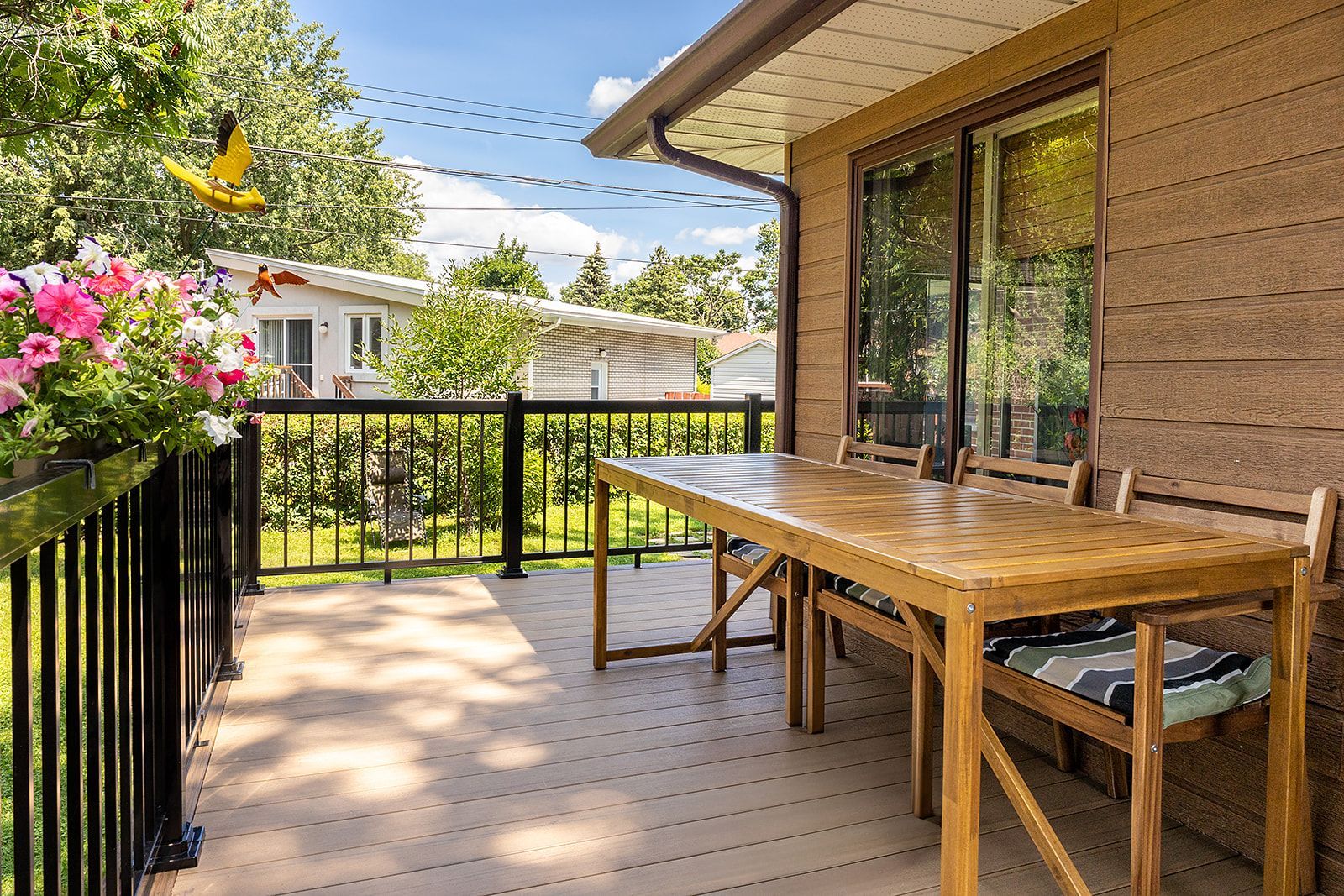 Terrasse en bois avec table et chaises, rambarde noire, jardinière, donnant sur le jardin et les maisons.