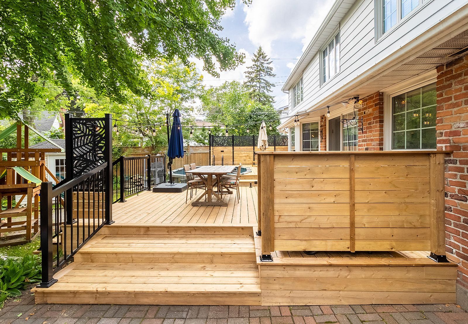 Terrasse en bois avec marches, rambarde noire et panneau décoratif, jouxtant une maison en briques.