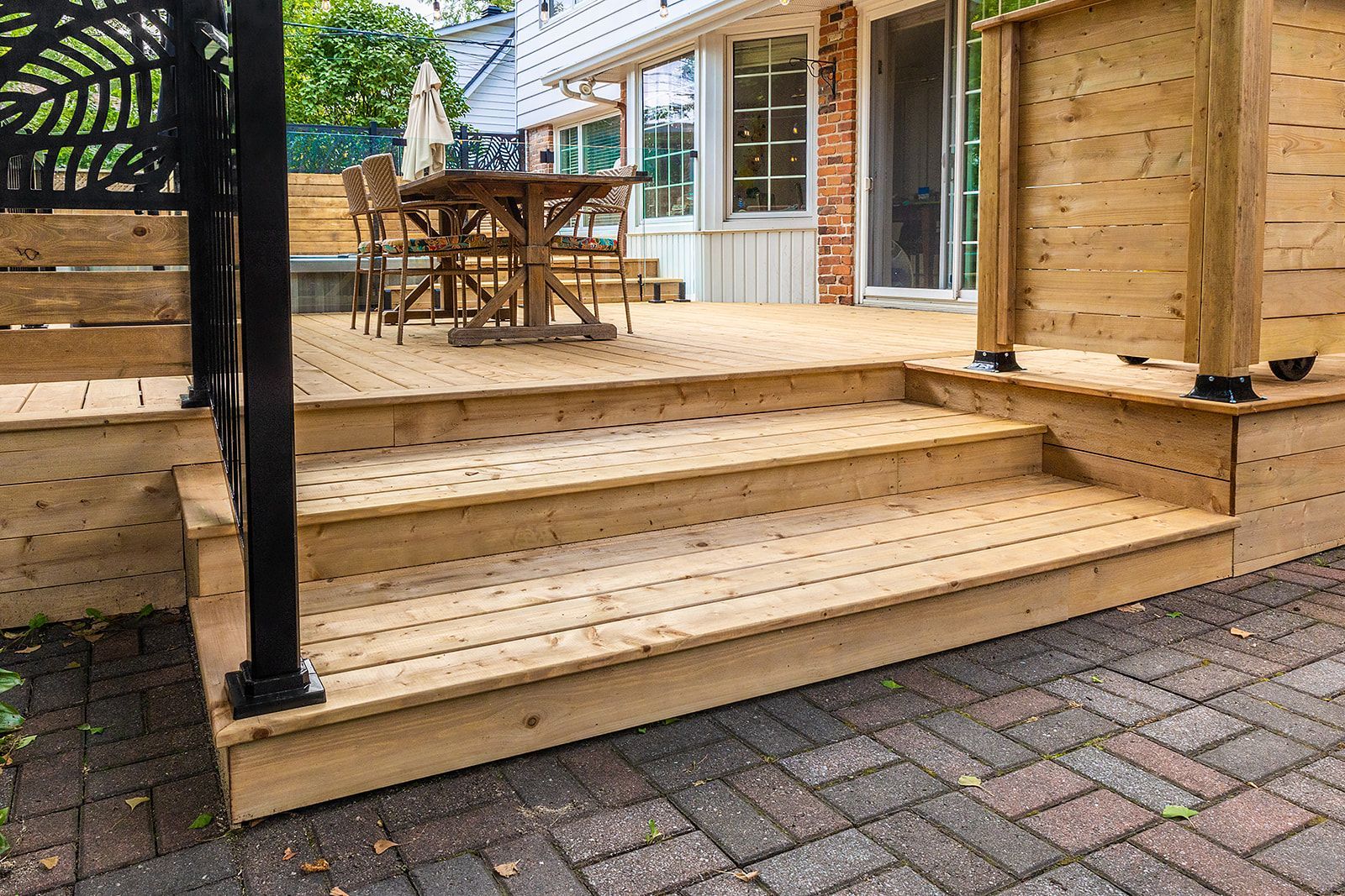 Terrasse en bois à trois marches menant à un patio avec une table et des chaises, près d'un patio en briques.