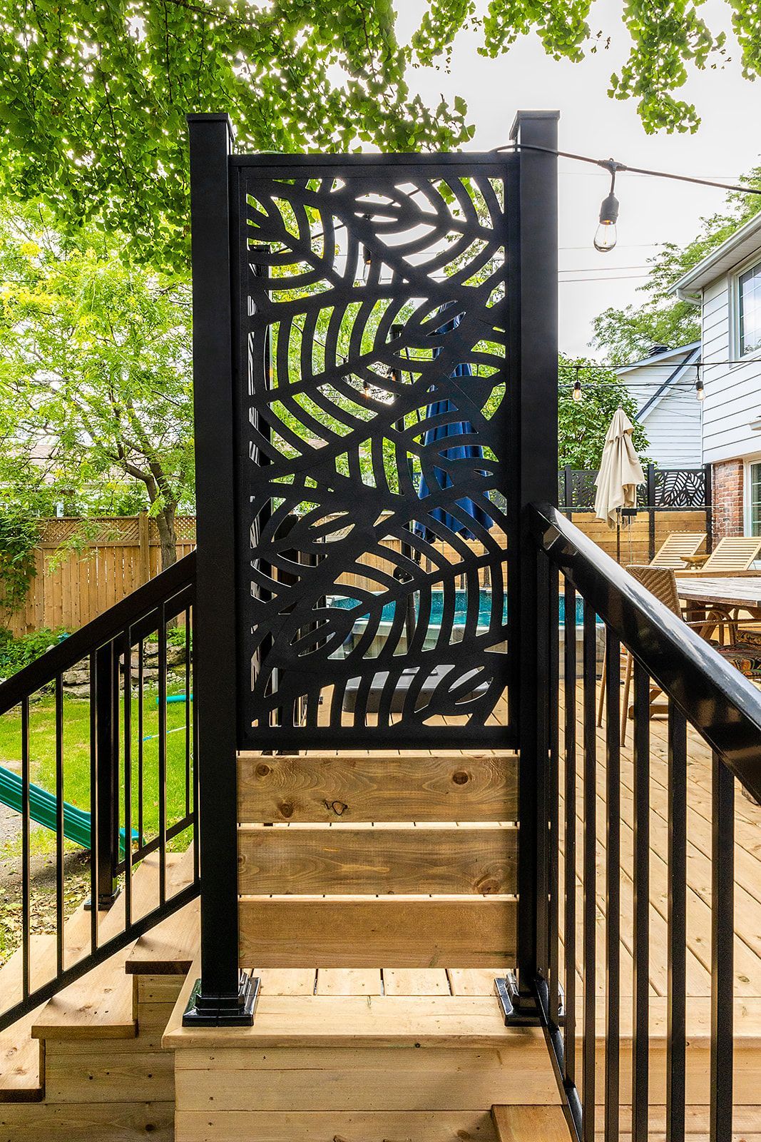 Paravent à motifs de feuilles noires sur les marches de la terrasse en bois. Rambarde noire. Jardin avec verdure et piscine.