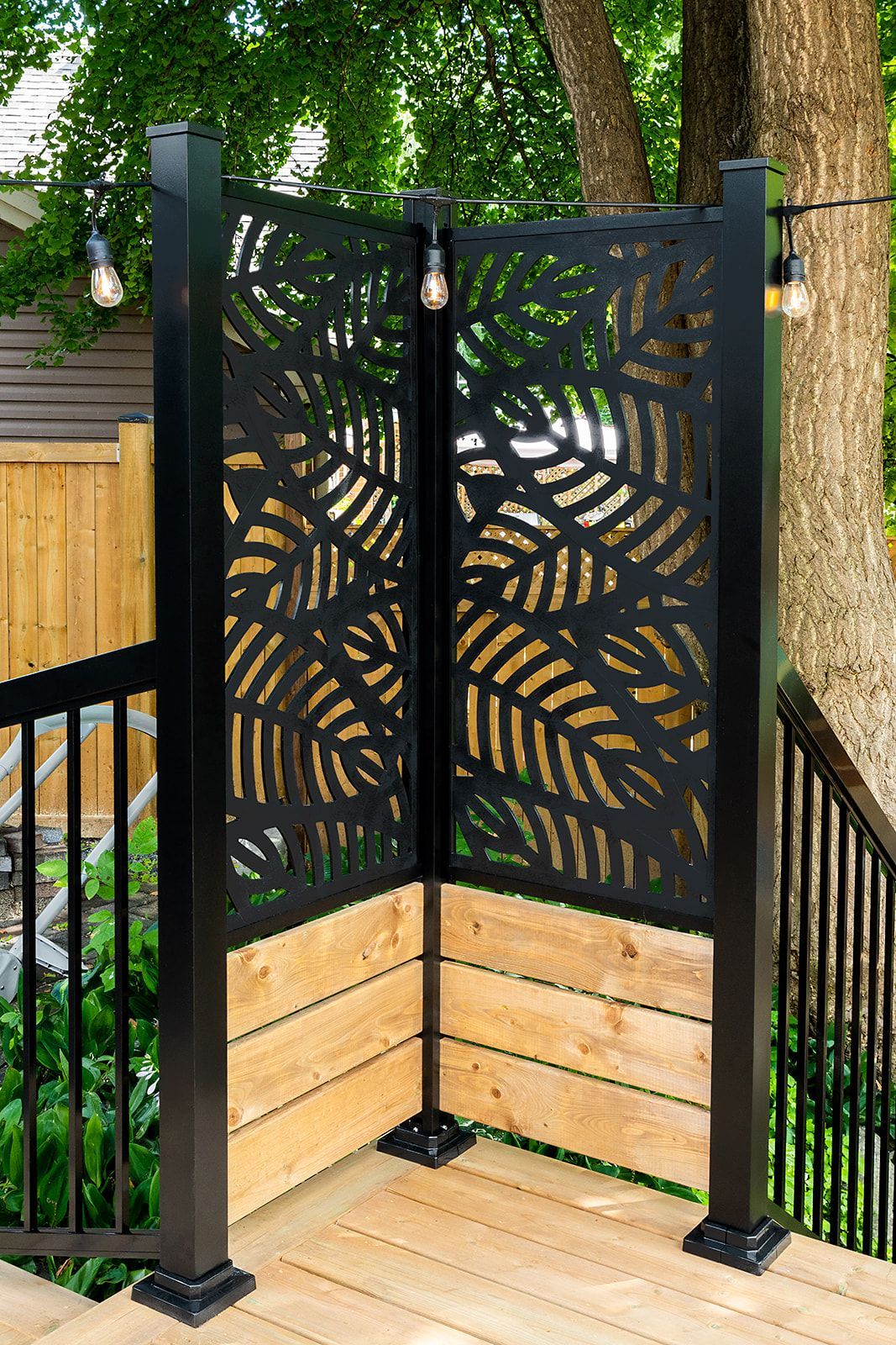 Paravent noir à motif de feuilles découpé au laser, installé sur une terrasse en bois et flanqué de poteaux noirs avec éclairage.