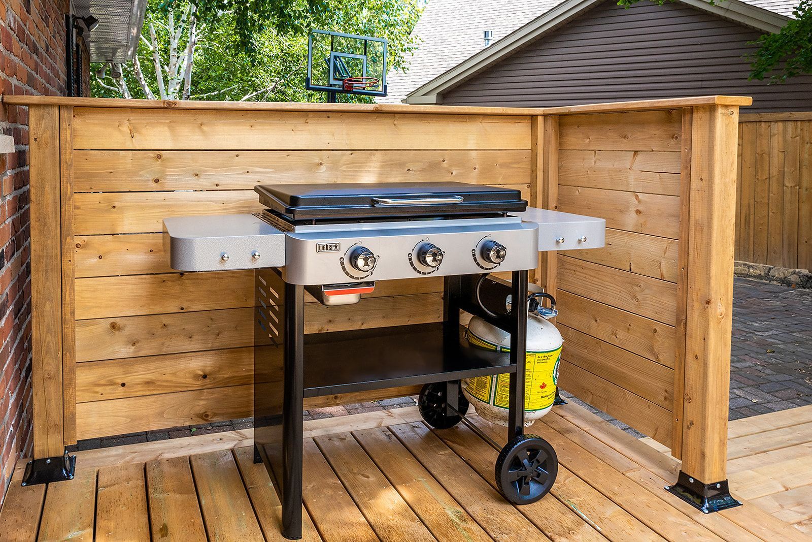 Barbecue sur une terrasse en bois, entourée d'une clôture en bois. Grille noire, accents gris et argentés, roues noires.
