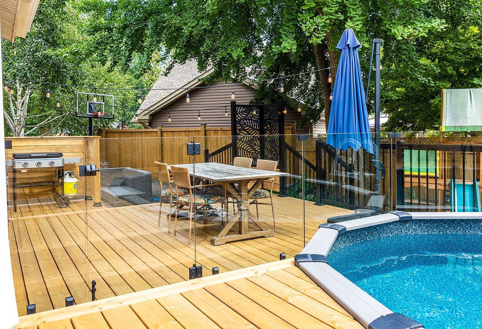 Terrasse en bois avec coin repas extérieur et piscine, entourée d'une clôture en bois et de verdure.