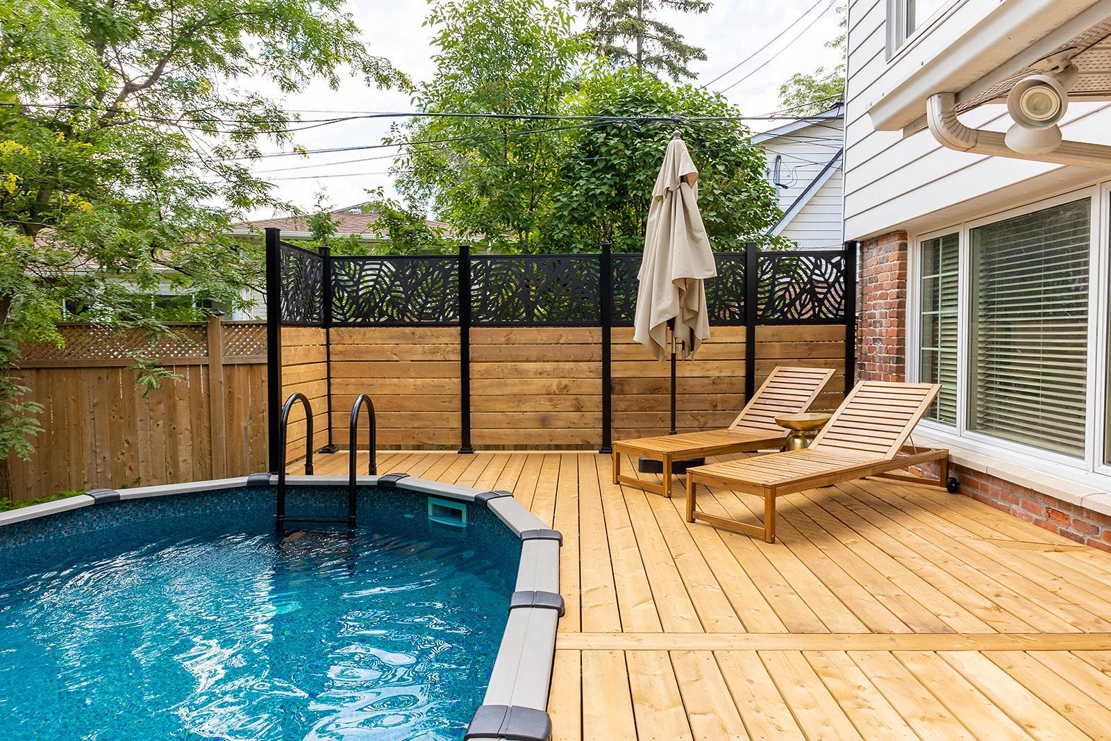 Jardin avec piscine, terrasse et chaises longues attenant à une maison. Clôture en arrière-plan, journée ensoleillée.