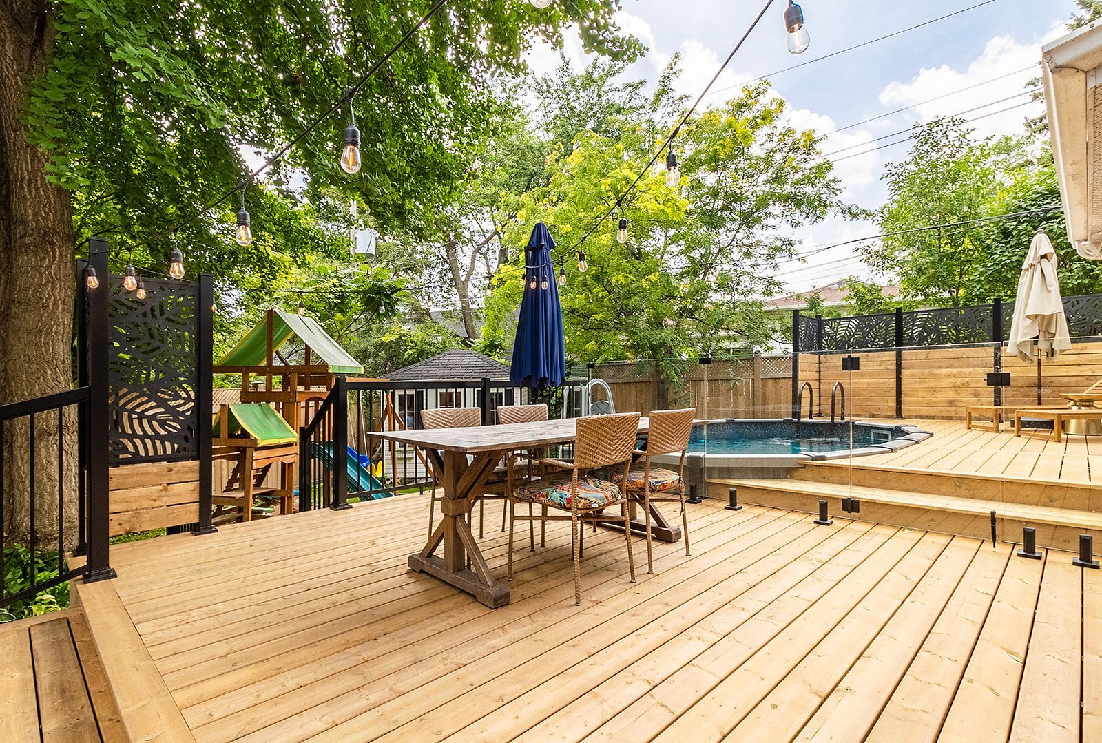 Terrasse en bois avec table à manger, chaises et petite piscine. Guirlandes lumineuses suspendues ; végétation luxuriante tout autour.