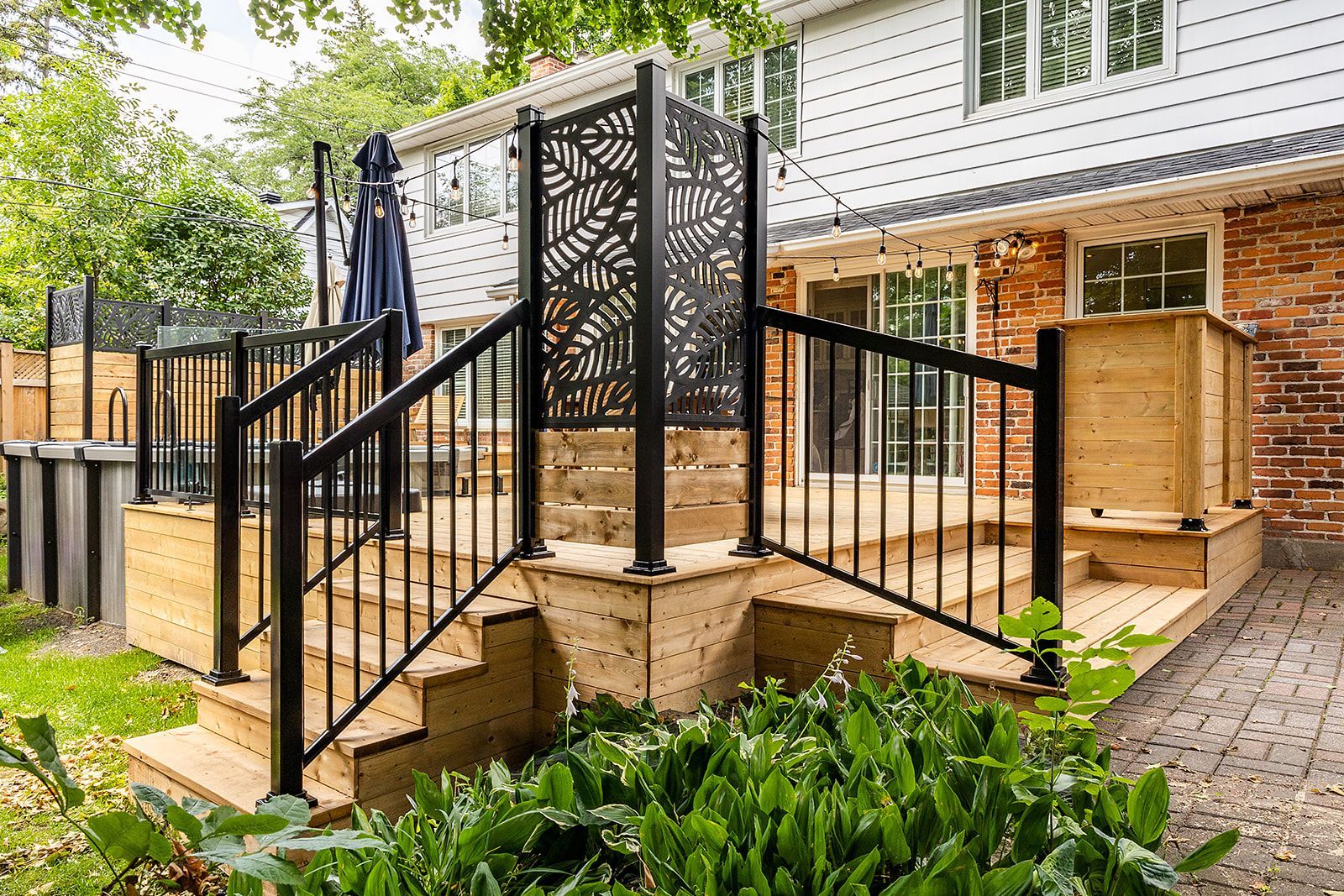 Terrasse en bois avec rambarde noire et panneau décoratif, jouxtant une maison en briques.