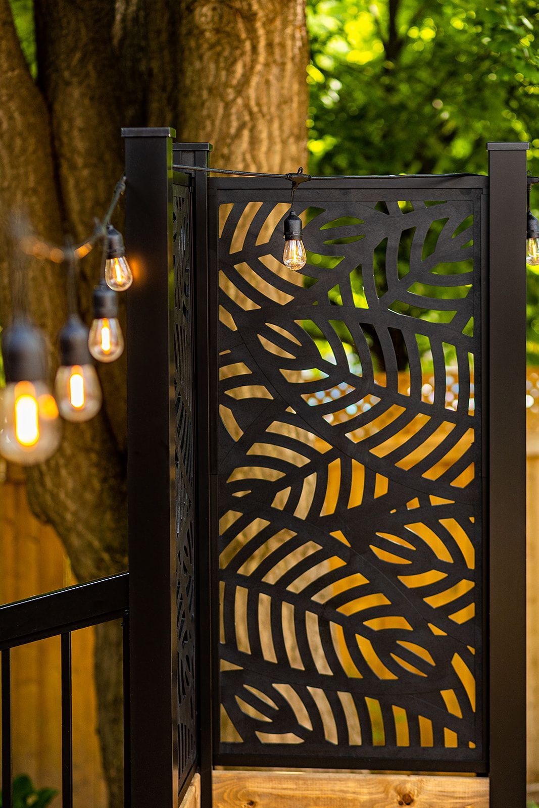 Paravent en métal noir à motifs de feuilles sur une terrasse en bois, avec guirlandes lumineuses et arbres en arrière-plan.