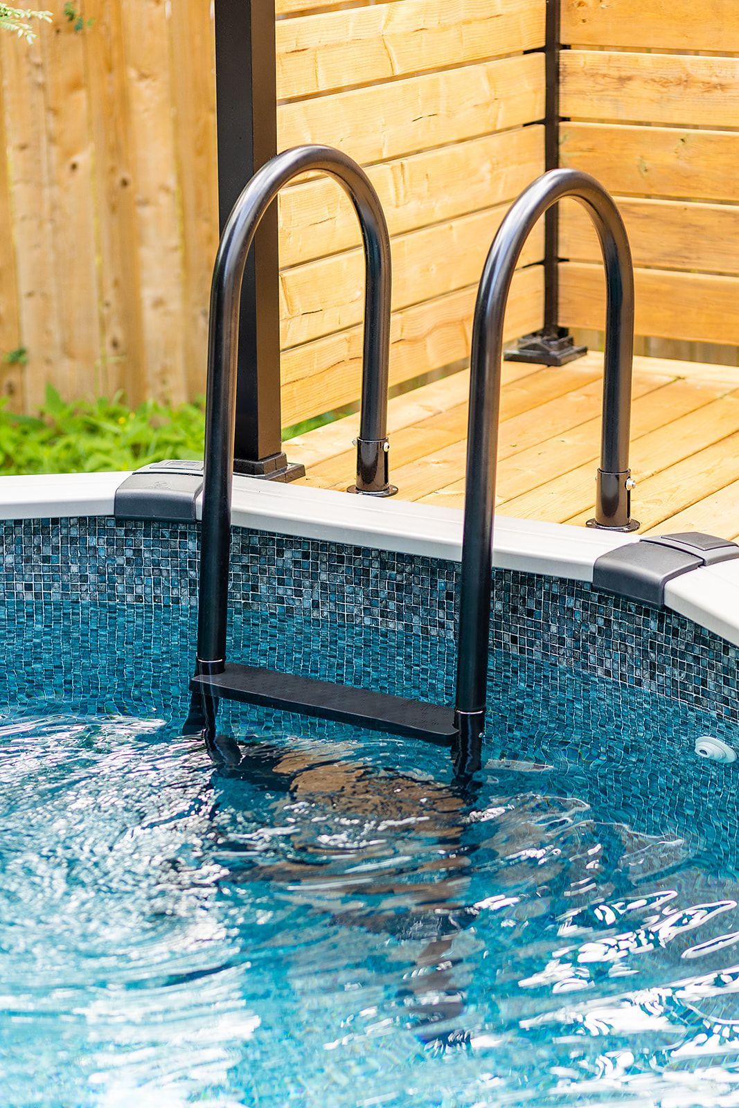 Échelle de piscine noire dans l'eau bleue, terrasse en bois et clôture en arrière-plan.