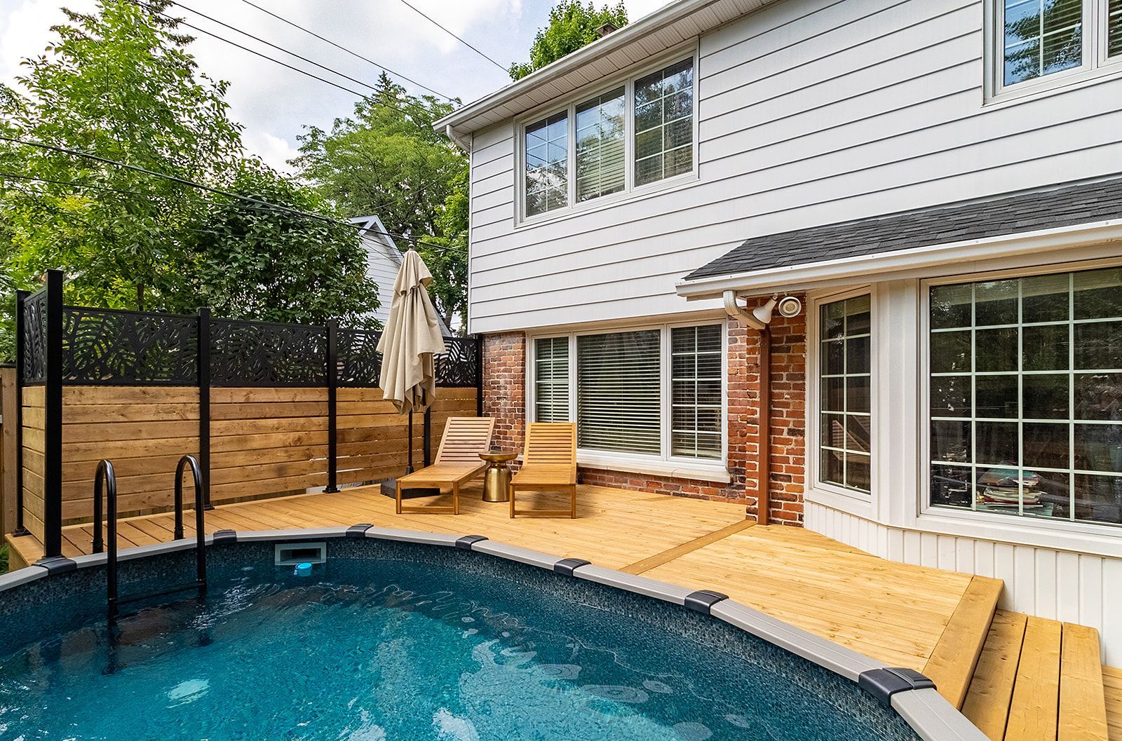 Cour arrière avec piscine hors sol, terrasse en bois, chaises longues et maison blanche à deux étages.