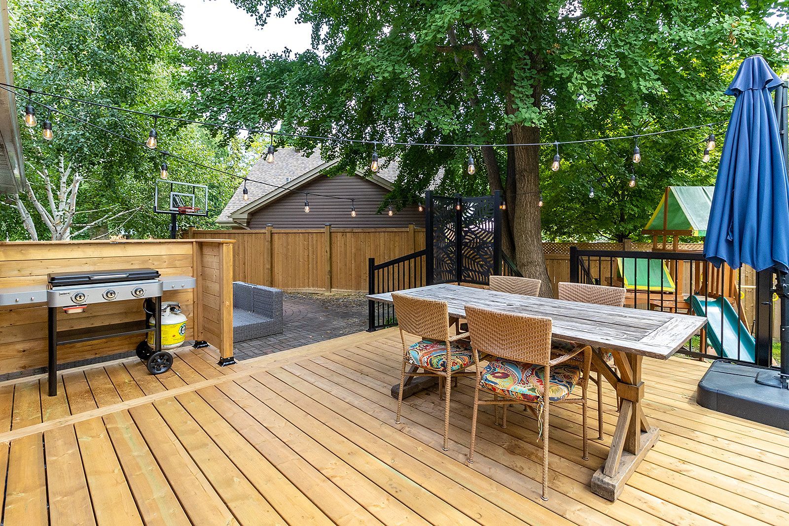 Terrasse en bois avec barbecue, table, chaises et parasol dans un jardin.