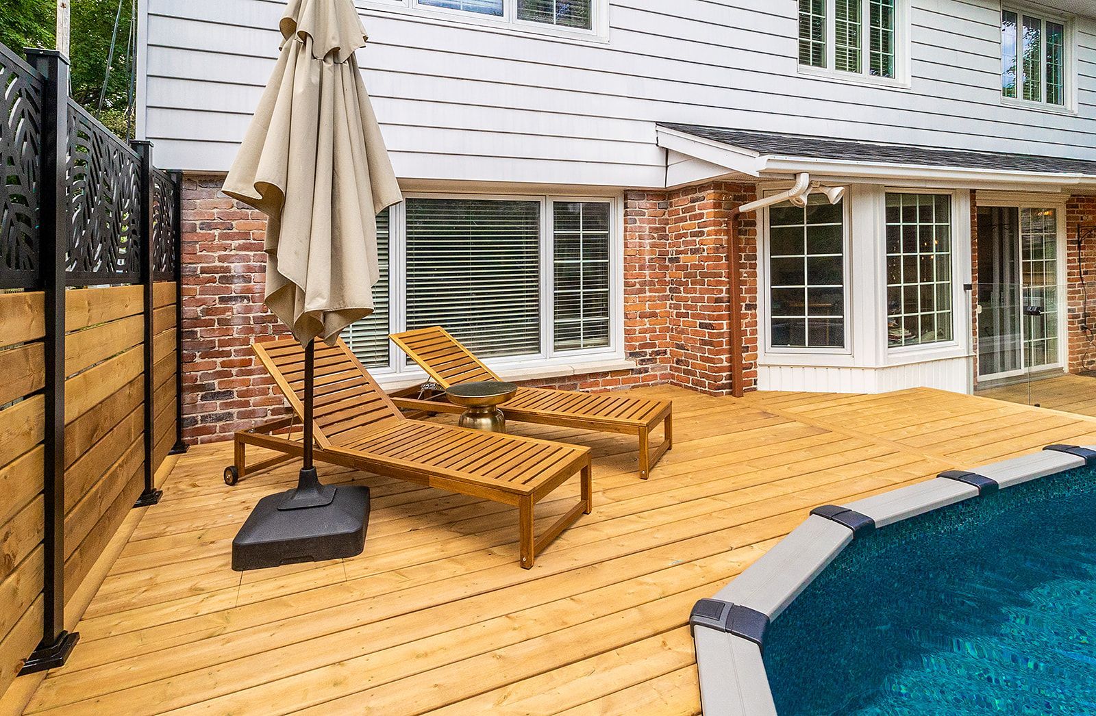 Terrasse en bois avec chaises longues, parasol et piscine à côté d'une maison en briques.