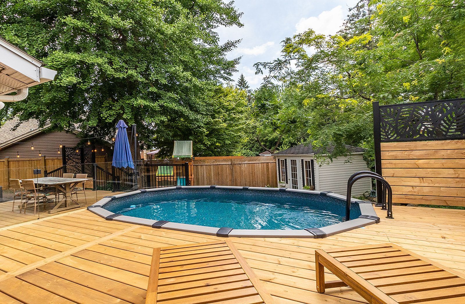 Piscine hors sol sur une terrasse en bois entourée d'arbres. Deux chaises longues sont au premier plan.