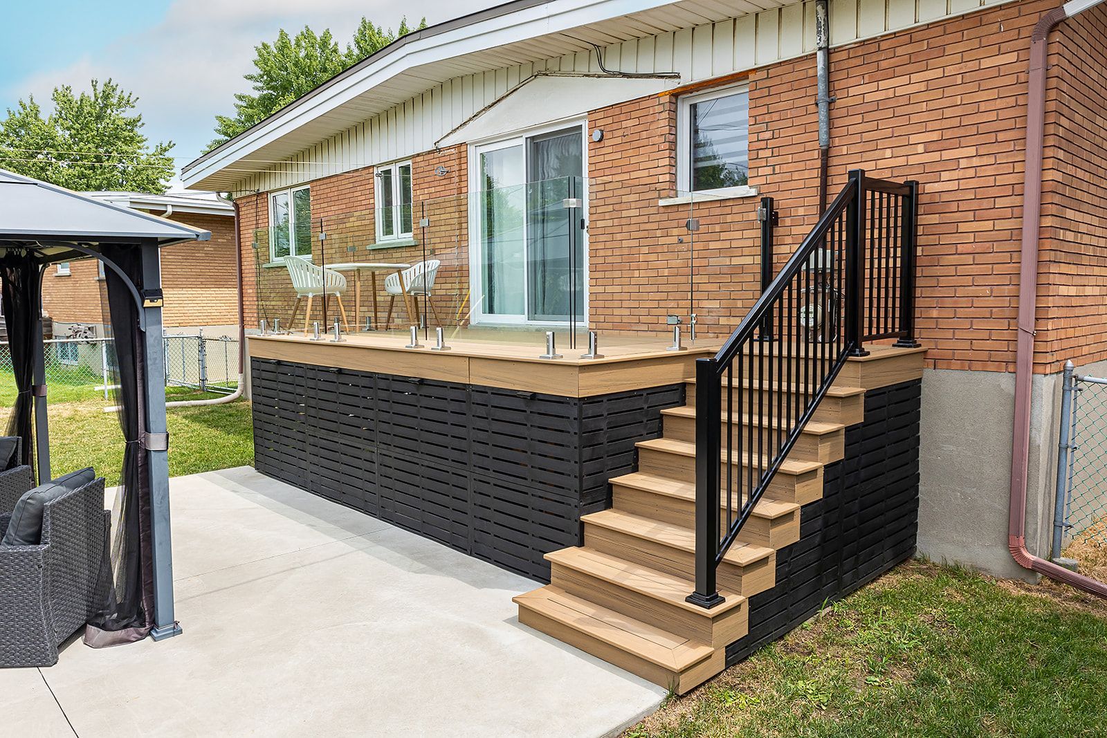 Terrasse arrière avec escalier, rambarde noire, mur de briques et patio en béton.