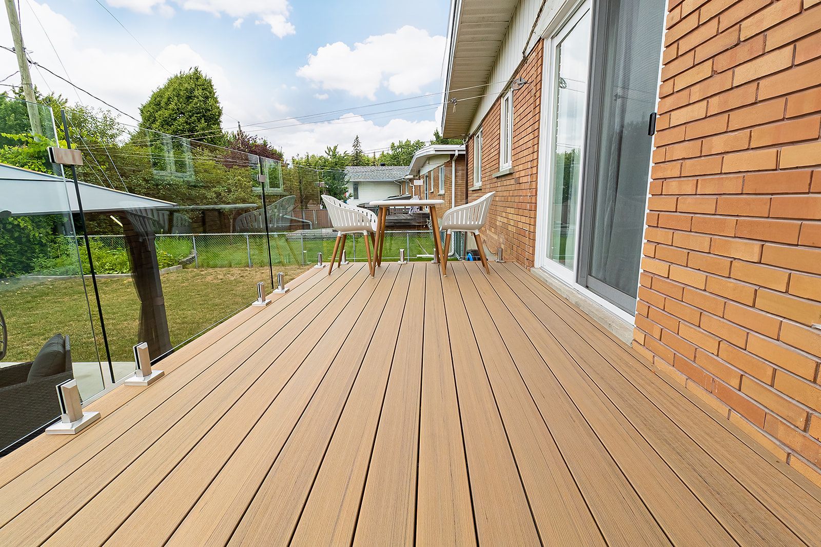 Une terrasse en bois prolonge un bâtiment en briques avec une rambarde en verre ; des chaises et une table sont visibles au loin.