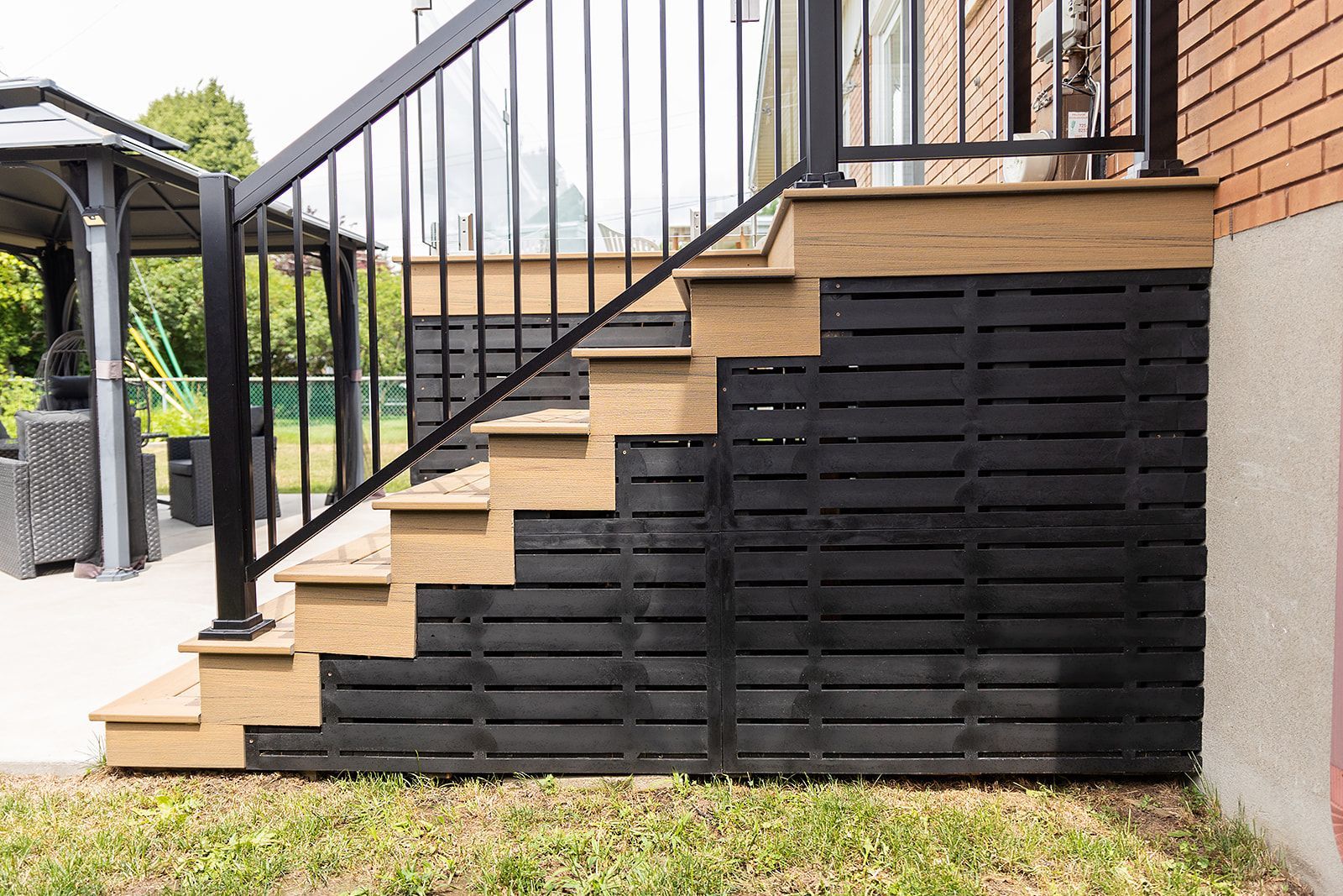 Escalier extérieur en bois avec rampe et plinthe noires, jouxtant un mur de briques et une pelouse.