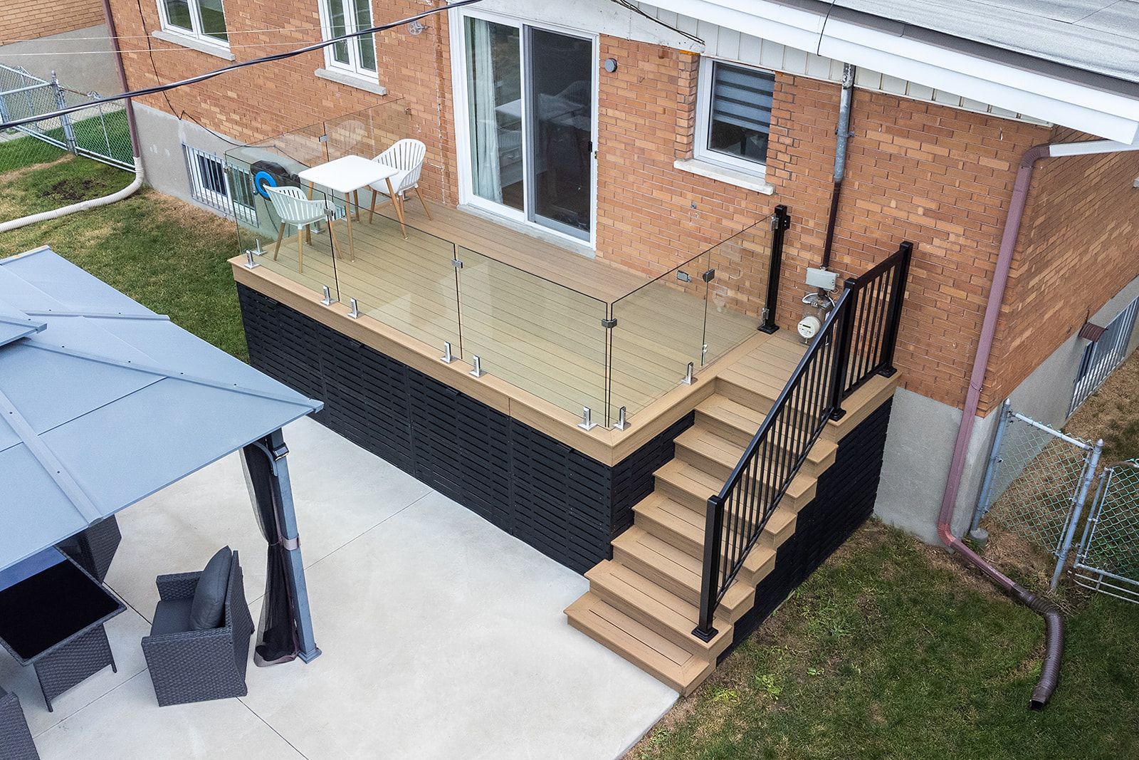 Terrasse avec balustrade en verre, escalier et gazebo jouxtant une maison en briques.