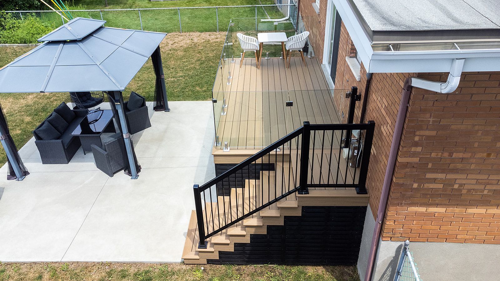 Terrasse avec escalier, rambarde noire et gazebo avec sièges sur un patio en béton à côté d'une maison en briques.