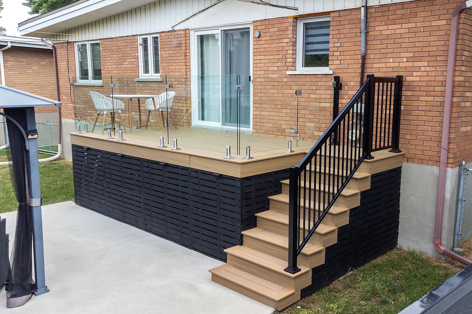 Une maison en briques avec une terrasse et un escalier en composite noir, des balustrades marron et une porte coulissante en verre.