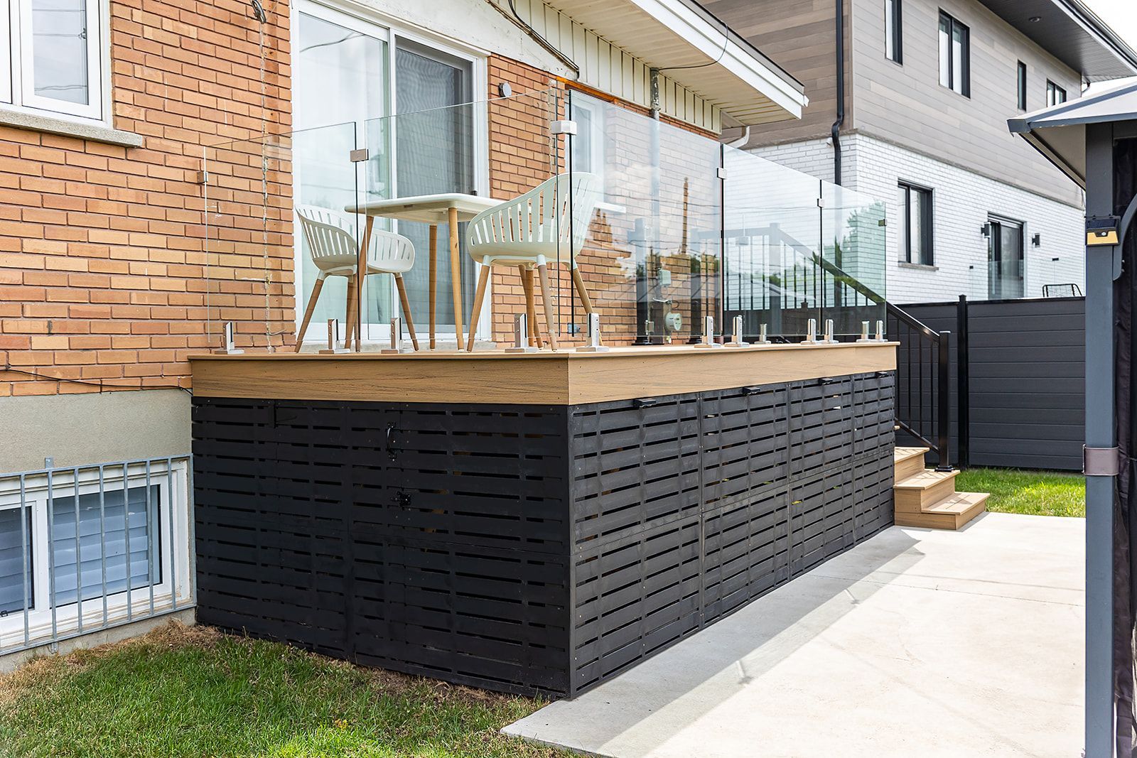 Terrasse arrière avec balustrade en verre, façade noire, petite table et chaises. Maison en briques en arrière-plan.