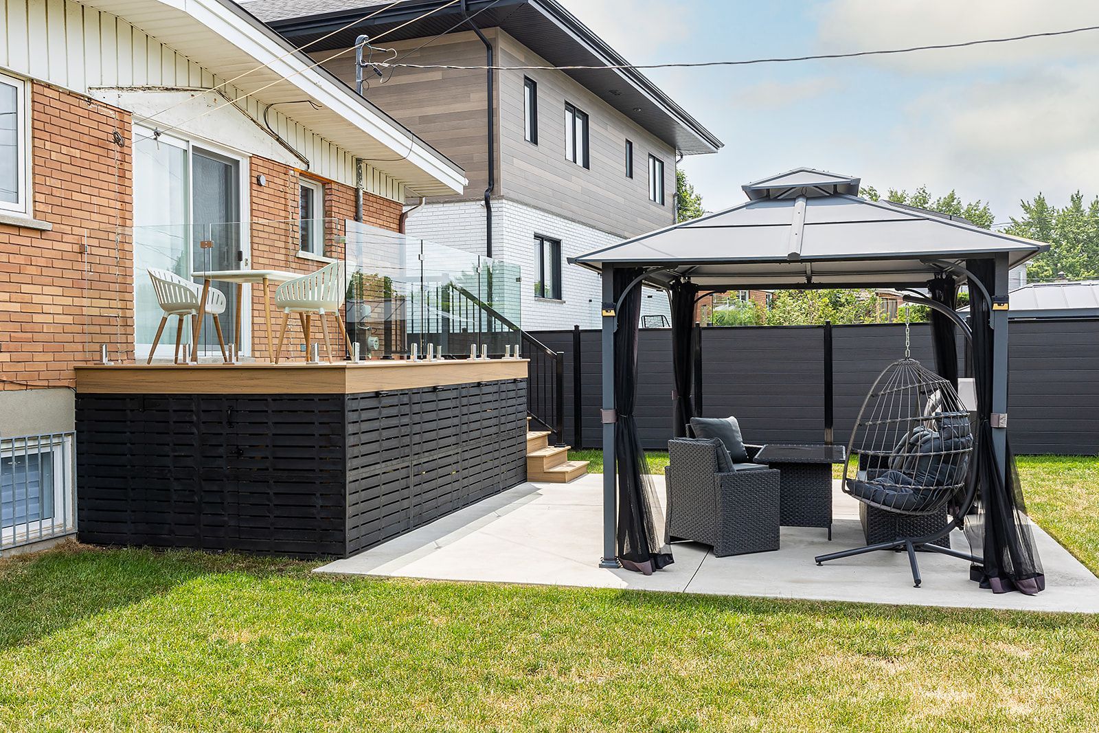 Terrasse arrière avec un kiosque, des sièges et une terrasse avec balustrade en verre, à côté d'une maison en briques.
