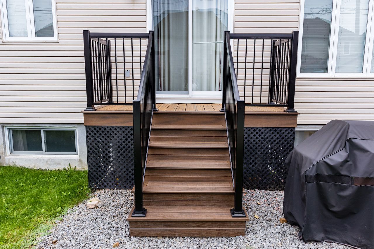 Terrasse et escalier en bois menant à une porte coulissante en verre, rambarde noire, bardage clair et sol en gravier gris.
