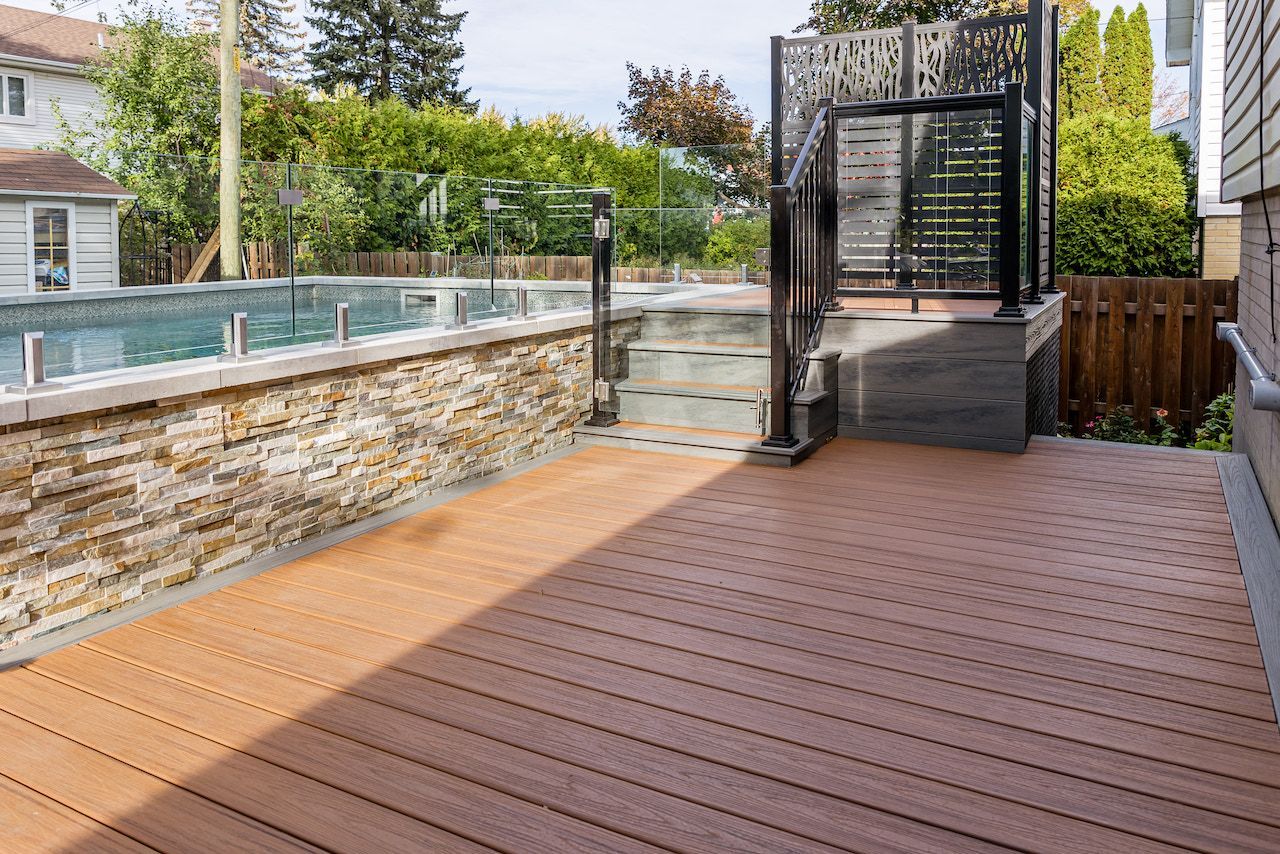 Terrasse en composite marron à côté d'une piscine avec un mur en pierre et une rambarde en métal noir.