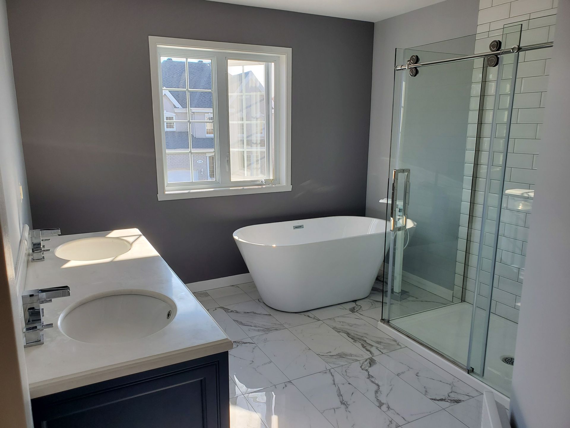 Salle de bains moderne avec baignoire blanche, sol en marbre, douche en verre, double vasque et fenêtre.