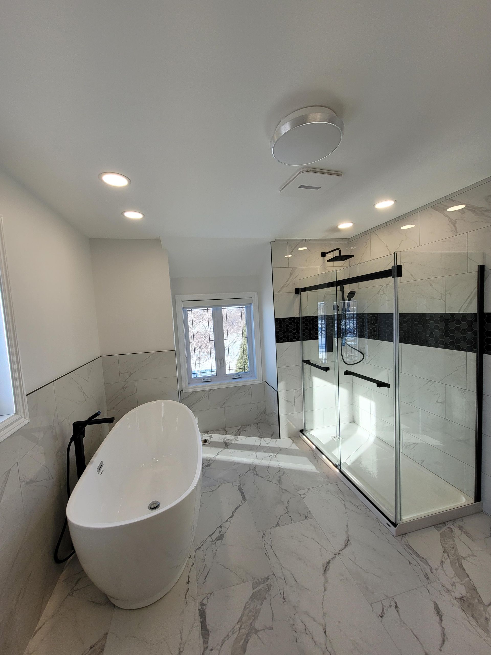 Salle de bains moderne avec baignoire îlot blanche, douche en verre, carrelage en marbre et éclairage encastré.