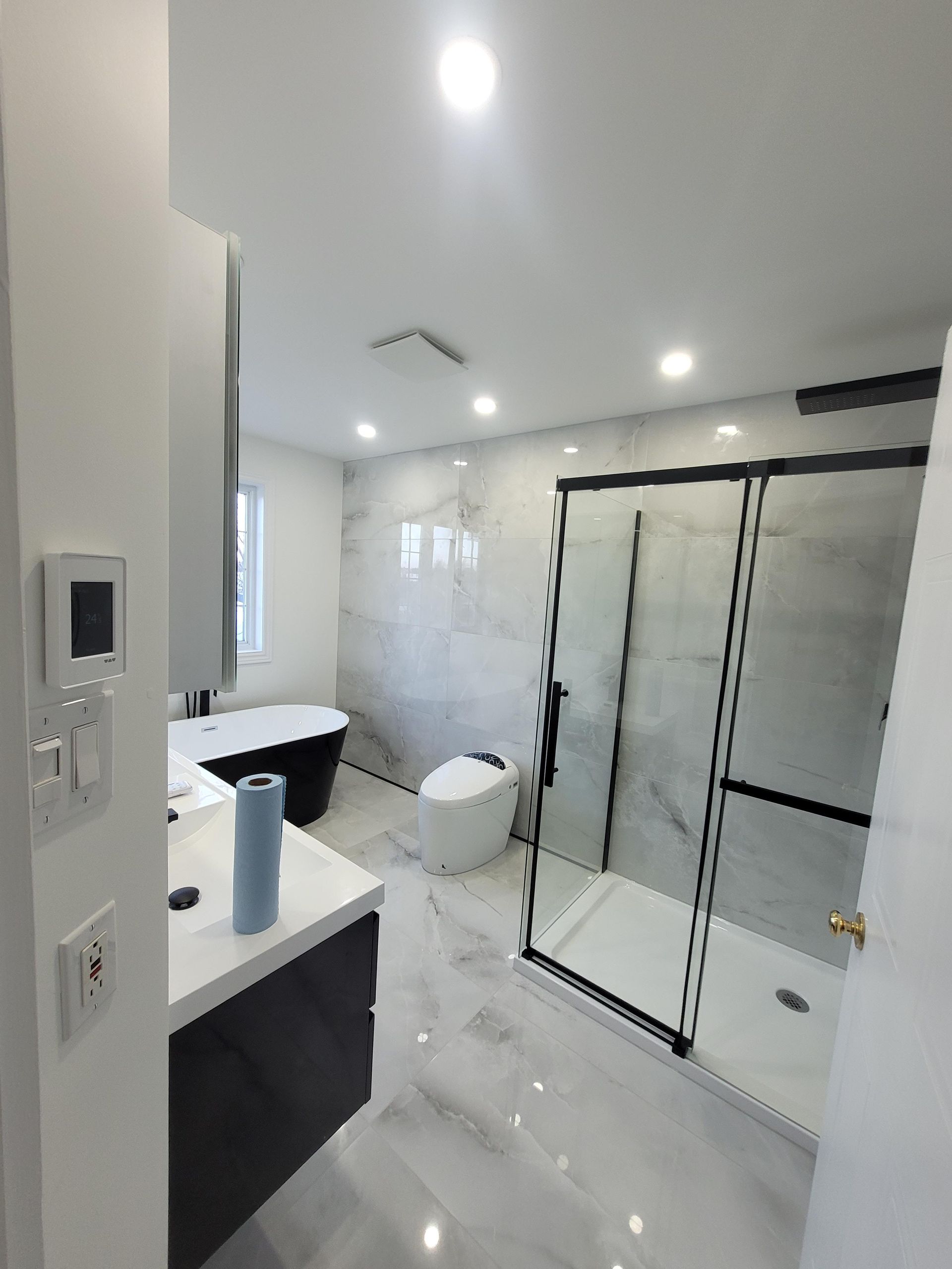 Salle de bains moderne avec murs et sol carrelés gris. Douche à cadre noir, meuble-lavabo noir et baignoire îlot.