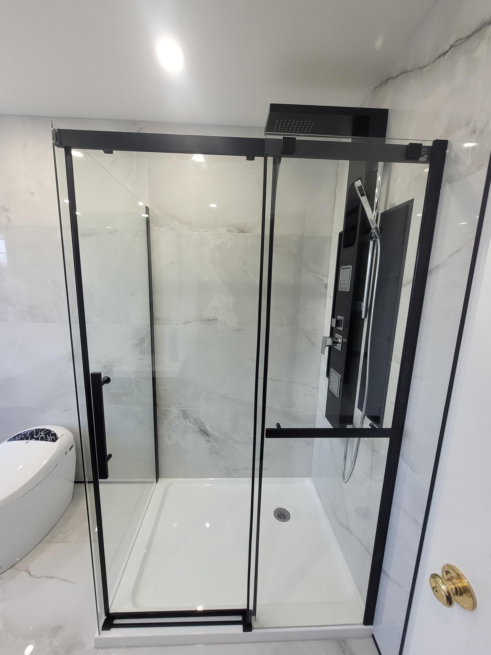 Cabine de douche en verre à cadre noir et base blanche, sur fond de carrelage imitation marbre. Salle de bains.