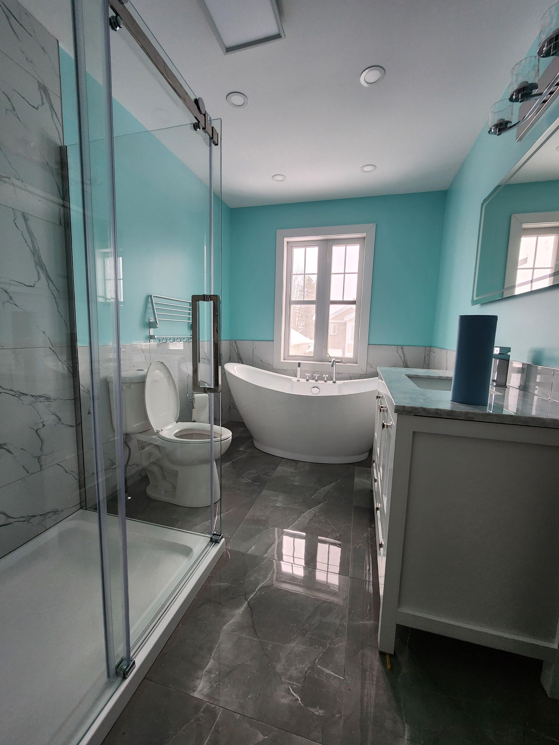 Salle de bains avec murs turquoise, carrelage gris, baignoire blanche, toilettes, meuble-lavabo et douche en verre.