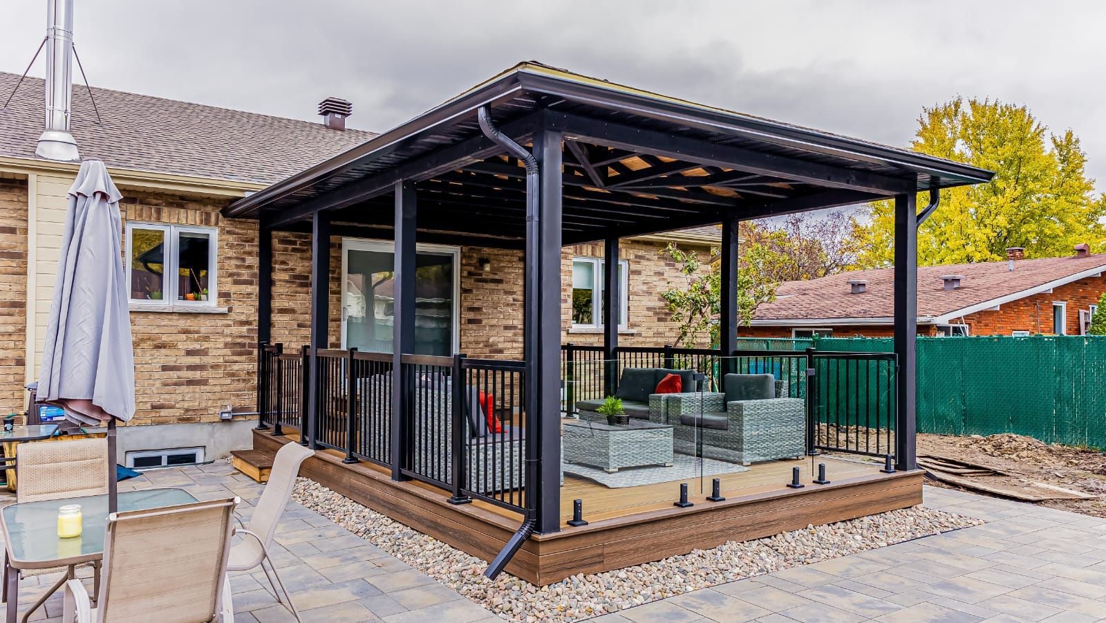 Patio couvert avec coin salon, attenant à une maison en briques. Structure noire avec rambarde et toit foncé.