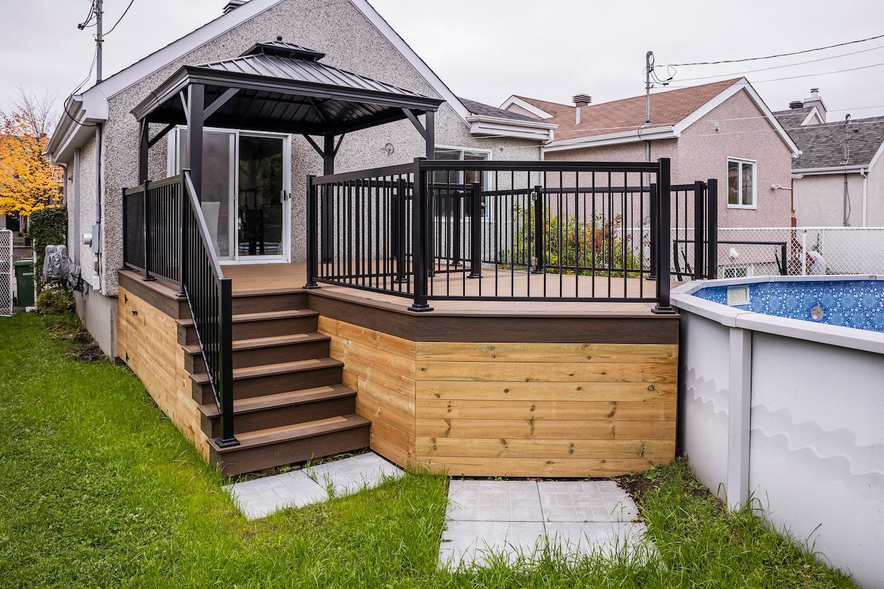 Terrasse en bois avec rambarde et escalier noirs, attenante à une maison avec piscine à proximité.