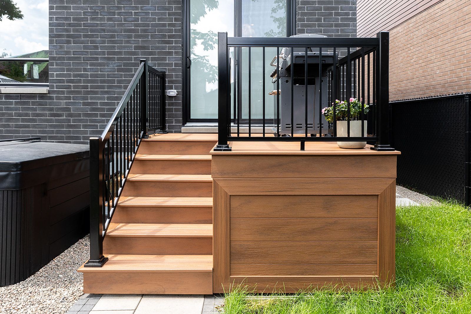 Terrasse en bois avec escalier, rambarde noire et rangements intégrés. Quelques marches mènent à une porte donnant sur le jardin.