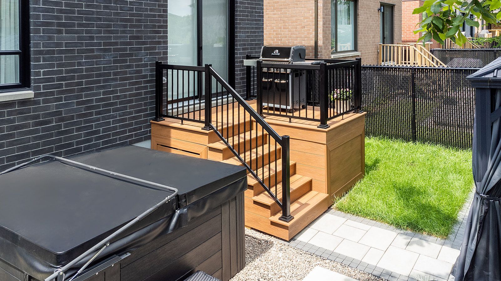 Jardin avec terrasse en bois surélevée, barbecue, escalier et jacuzzi. Mur de briques noires et pelouse verte.