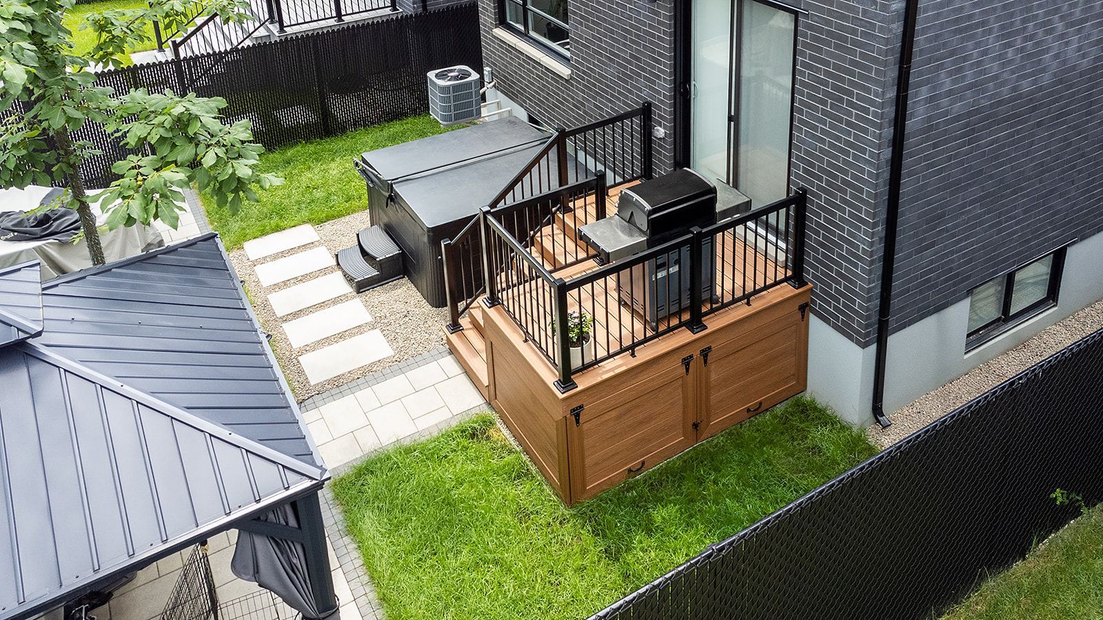 Terrasse arrière avec jacuzzi, allée en pierre et barbecue. Maison en briques noires, pelouse verte et clôture noire.