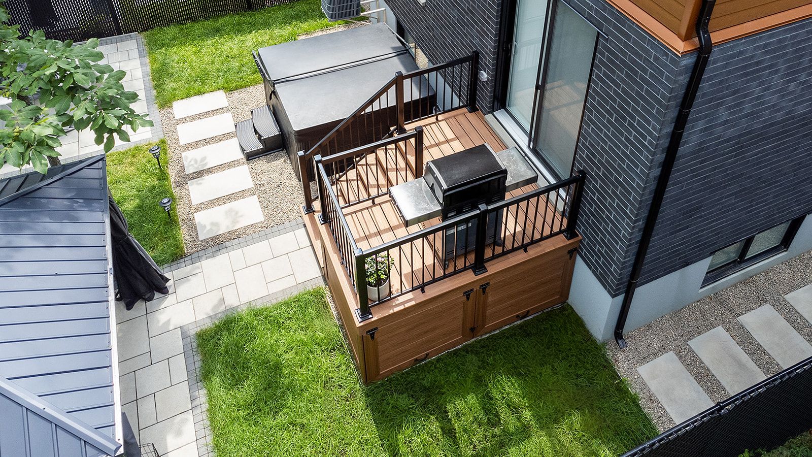 Terrasse en bois surélevée avec barbecue, accessible depuis un bâtiment sombre muni d'une porte. Pelouse et chemin de pierre entourent la terrasse.
