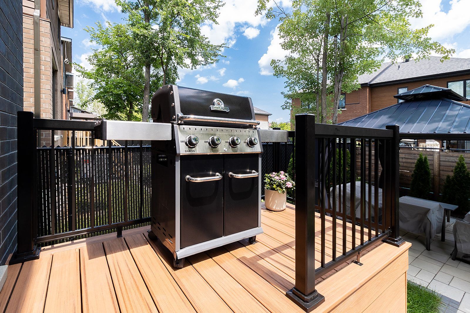 Barbecue à gaz noir sur une terrasse en bois avec rambardes noires ; arbres et kiosque à l'arrière-plan.