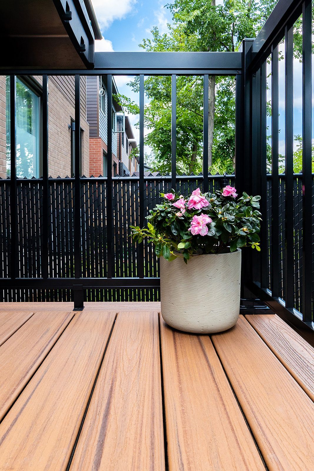 Terrasse en bois avec une plante fleurie rose en pot, sur fond de rambarde noire et de verdure.