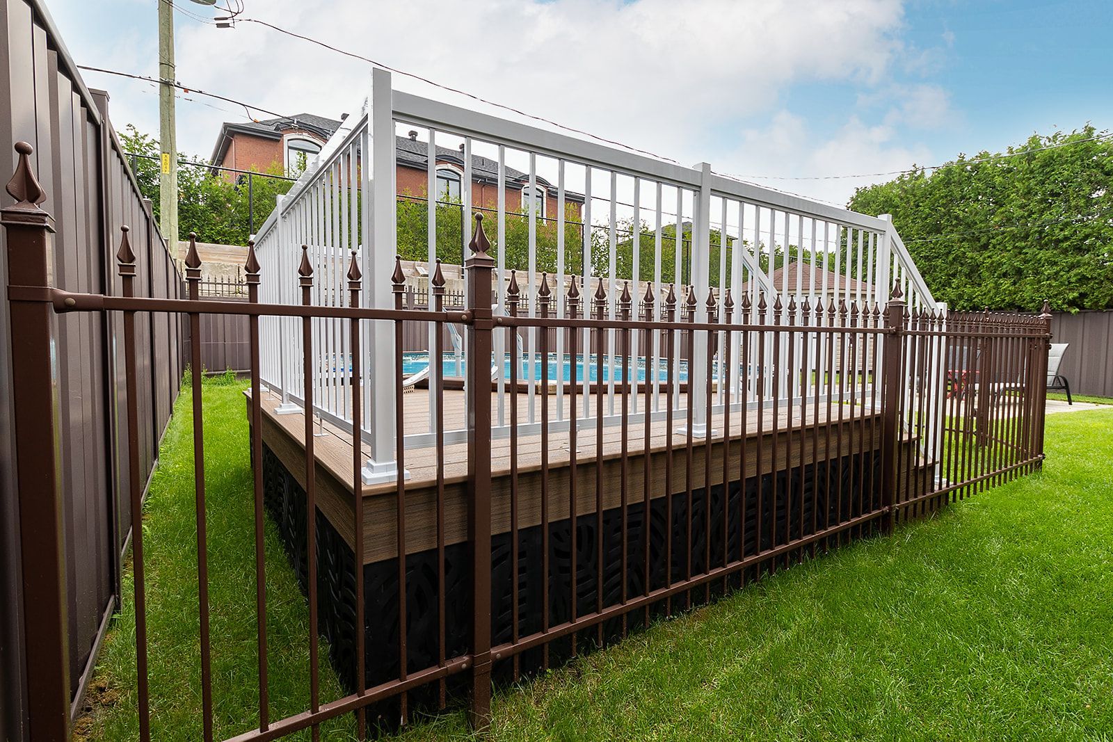 Une terrasse clôturée de brun avec une rambarde blanche entoure une petite piscine, sur une pelouse verte, sous un ciel bleu.